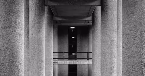 Black-and-white photograph of a brutalist concrete colonnade — vertical columns crossed at right angles by a horizontal beam and railing, two perpendicular axes meeting in a dark structural void.